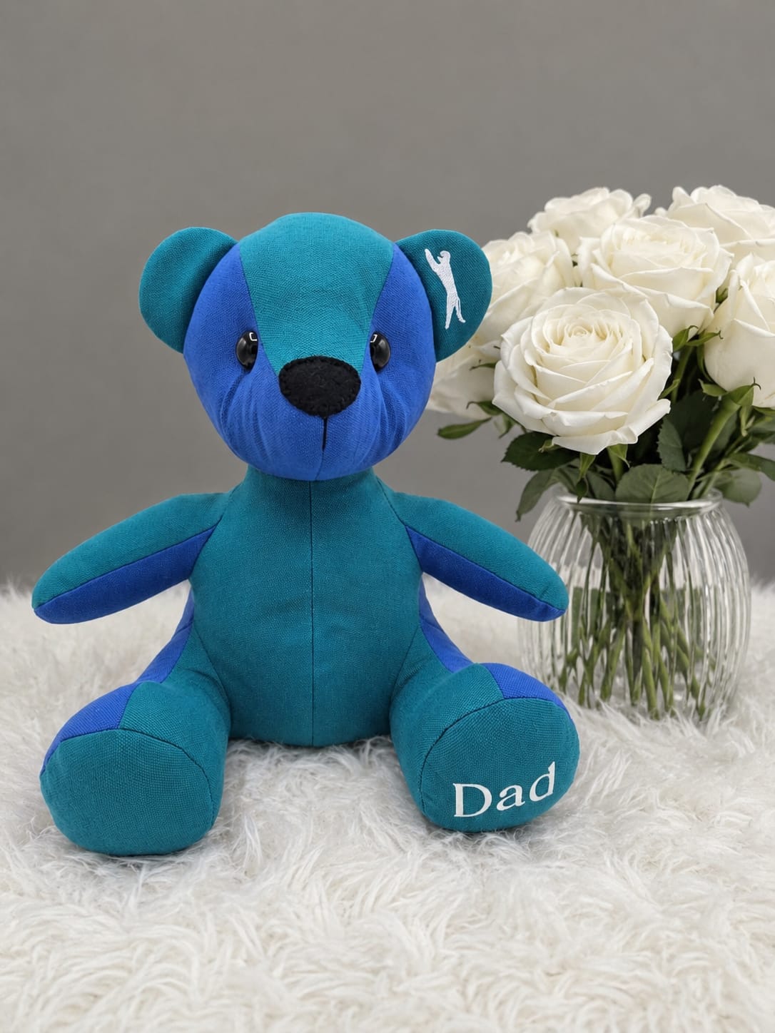 Blue and teal standard memory bear with Dad embroidered on its paw, beside white roses